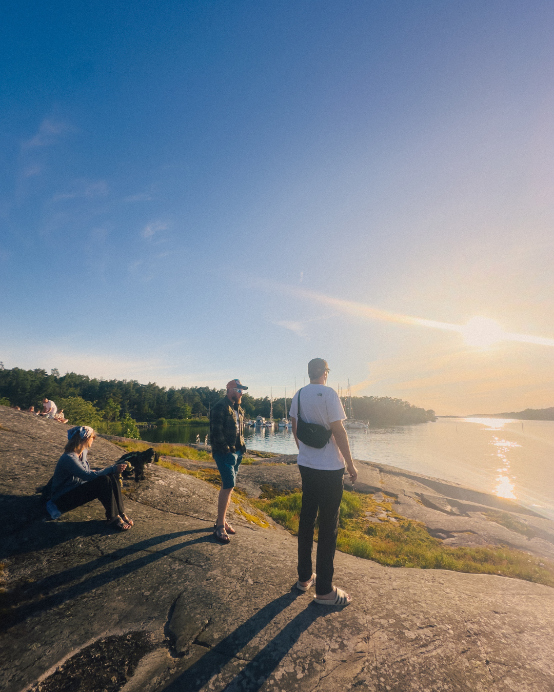 Ystävät istuvat kallioilla Gullkronassa auringonlaskun aikaan – Gullkronan satama ja purjeveneet näkyvät taustalla